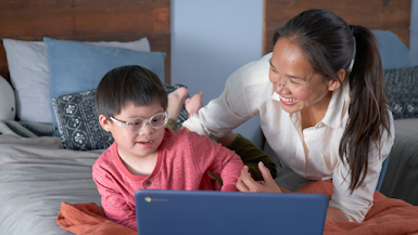 Un niño con camisa roja de manga larga y anteojos gruesos está recostado en una cama mirando una computadora portátil Chromebook, su madre está recostada a su lado sonriendo