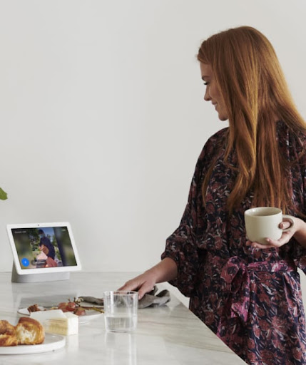 A woman drinking coffee looks at Nest Hub display on a table.