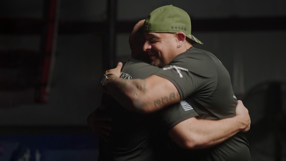 Two men wearing veterans gear and an camouflage-printed cap, hugging