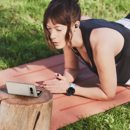 En person med et Wear OS-smart-ur og øretelefoner, der træner på en yogamåtte og kigger på en foldbar Android-telefon.