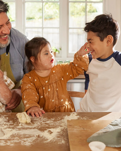 The daughter and her older brother play with ingredients as her father smiles and looks on.