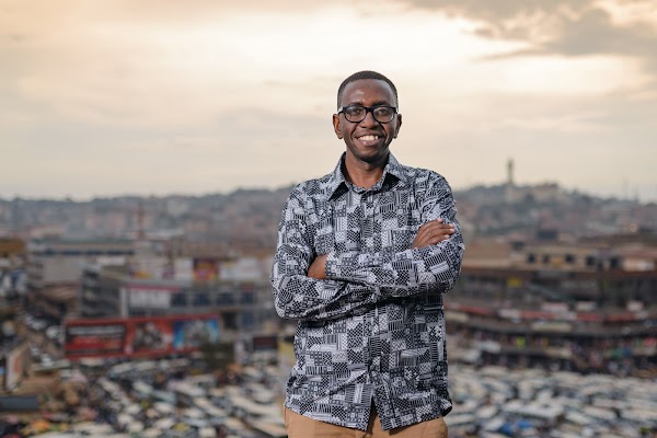 A man wearing glasses and a camouflage long sleeve shirt crosses his arms smiling against a background of a city