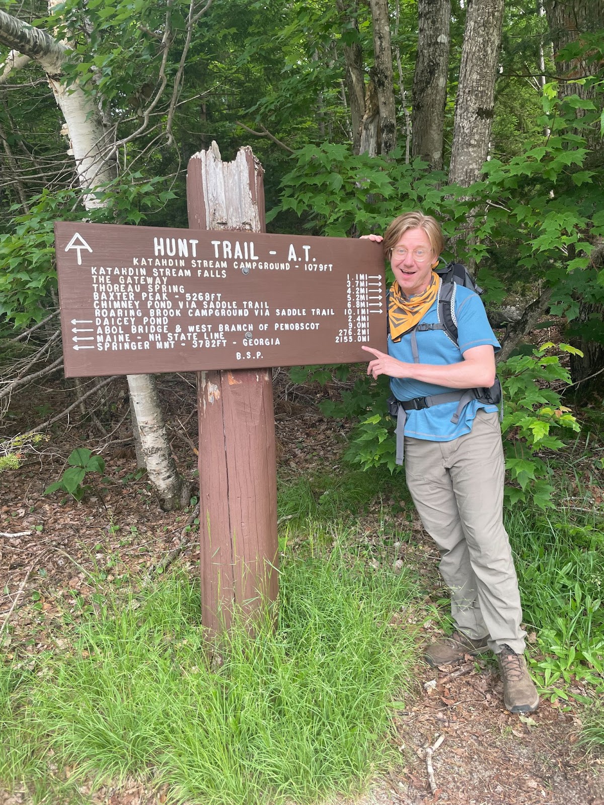 Photograph of a man standing in a forest next to a brown trailhead sign