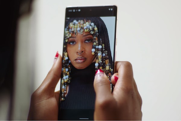 Someone with red tipped nails holds a smartphone. On the screen is a young woman with black braids and a black top facing the camera. Clear, white, black, and yellow beads adorn the ends of her braids.