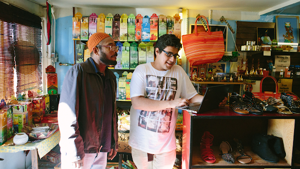 A boy smiles and stands next to a shop-owner in a store while they look at a laptop together