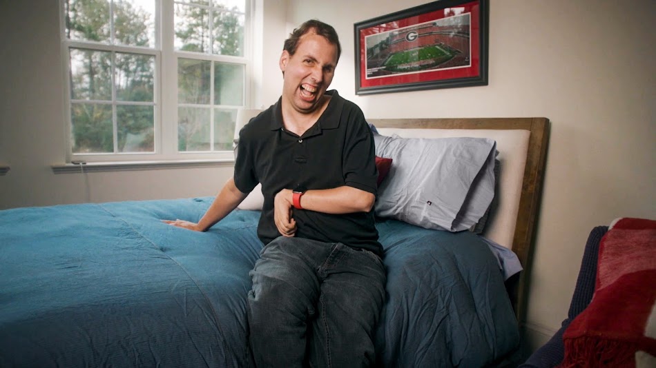 Person in black collared shirt sitting on bed smiling in front of a window.