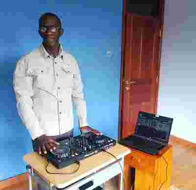 A photo of John, a tall young man, standing behind DJ turntables against a blue wall.