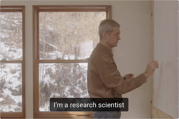 Dimitri Kanevsky, a fair skinned, white haired man, stands drawing on a whiteboard. Captions on the image show what Dimitri is saying- “I’m a research scientist.”