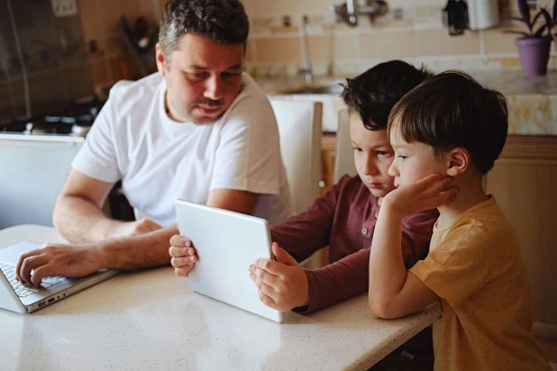 A father is overseeing his two sons as they use their tablet to access the internet for online learning