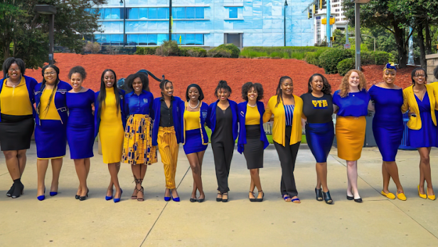 A group of 16 Black women wearing blue and yellow, standing in a line and smiling at the camera.