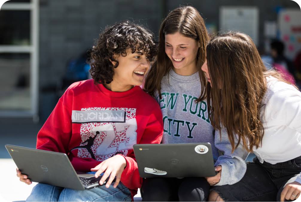 Three students smile as they work on two laptops outside.