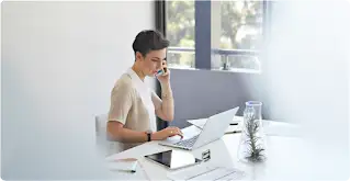 A short haired woman sits at a desk and talks on a smartphone with a laptop and tablet in front of her.