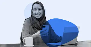 Una donna che indossa un hijab sorride mentre utilizza uno smartphone e un laptop.