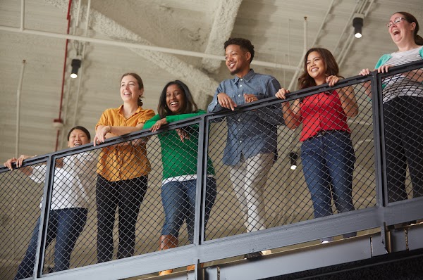 A group of happy Google employees in brightly colored shirts look down from the railing of a workplace stairwell