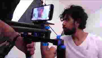 A photo of a movie being shot with an Android device and a device used to hold the camera steady. An arm holds the filming device while a young man appears in front of the camera.