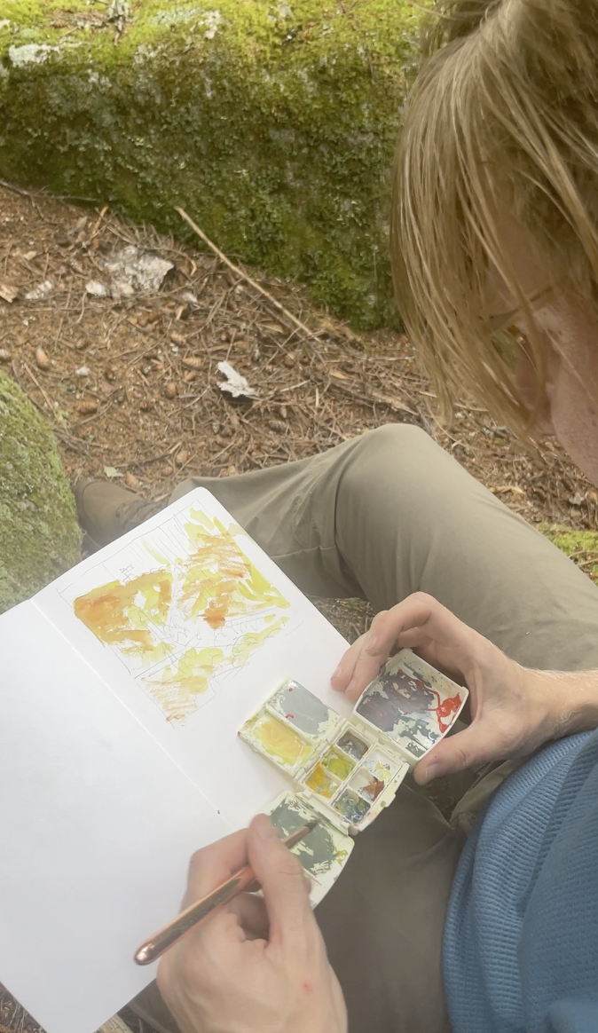 Photograph of a notebook with a work in progress painting resting on a person's leg in an outdoor setting