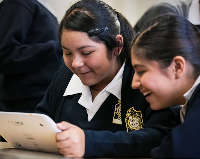 Two girls in school uniform smiling, one of them holding a tablet device