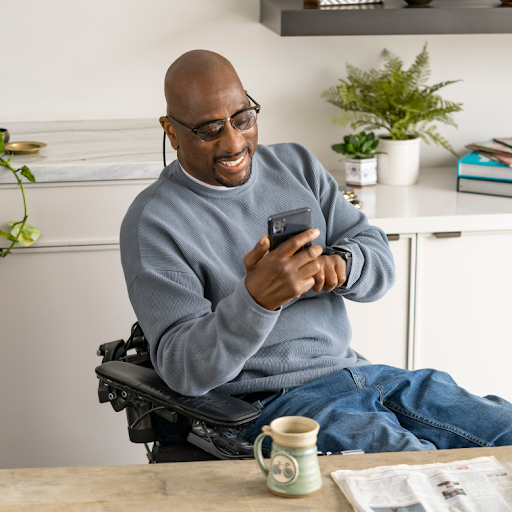 Un hombre afrodescendiente sentado en una silla de ruedas le sonríe a su teléfono