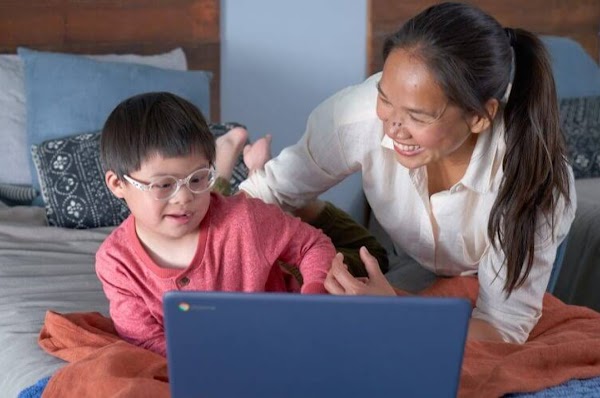 A young boy with glasses uses a laptop on a bed with his mom