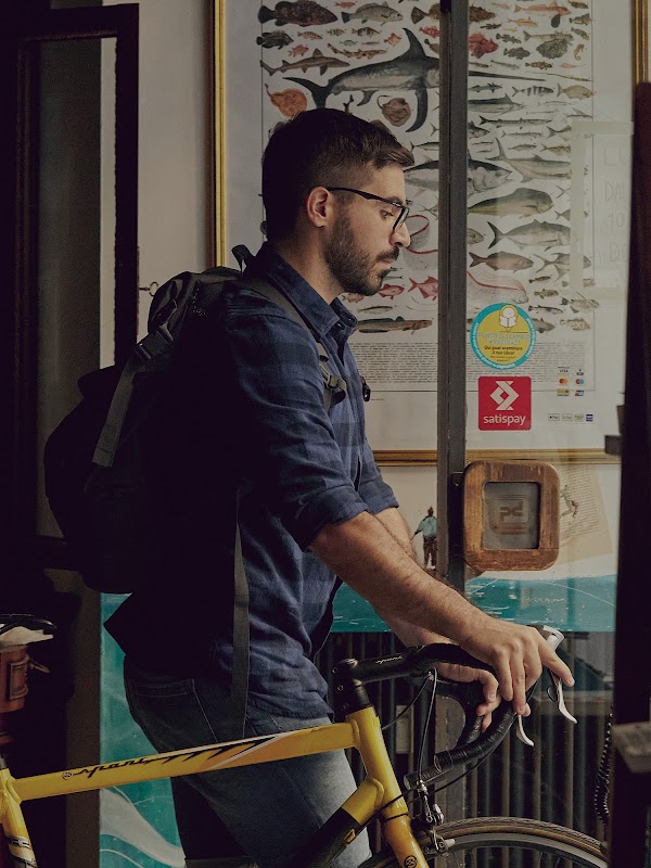 Mattia Garavaglia esce dalla sua bici gialla dalla sua libreria. Indossa occhiali e uno zaino.
