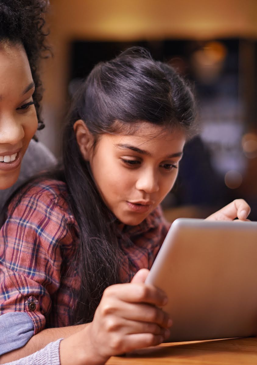 Una mujer sonriente y una niña curiosa miran juntas la pantalla de una tablet.