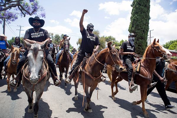 Randy y Los Vaqueros de Compton a caballo encabezando a millares de personas por las calles de Compton, luciendo artículos promocionales de Los Vaqueros de Compton, sombreros de vaquero y puños en alto.