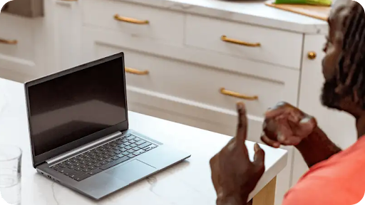 A Black man communicates in sign language to a person off camera at home. An open Chromebook is in front of him.