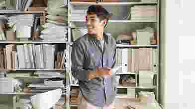 A younger man with dark hair, wearing a casual button-up and khakis, stands up from his desk, smiling and looking to the side. He holds a mobile phone. Shelves of papers and binders stand behind him.