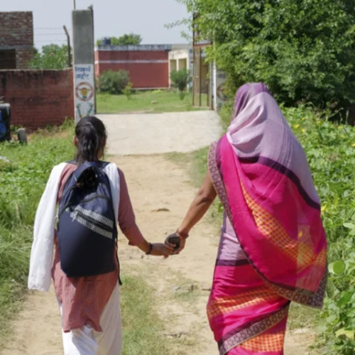 Two women, one carrying a backpack and the other dressed in a pink and purple sari, walk hand-in-hand down a pathway toward a building, with greenery on either side