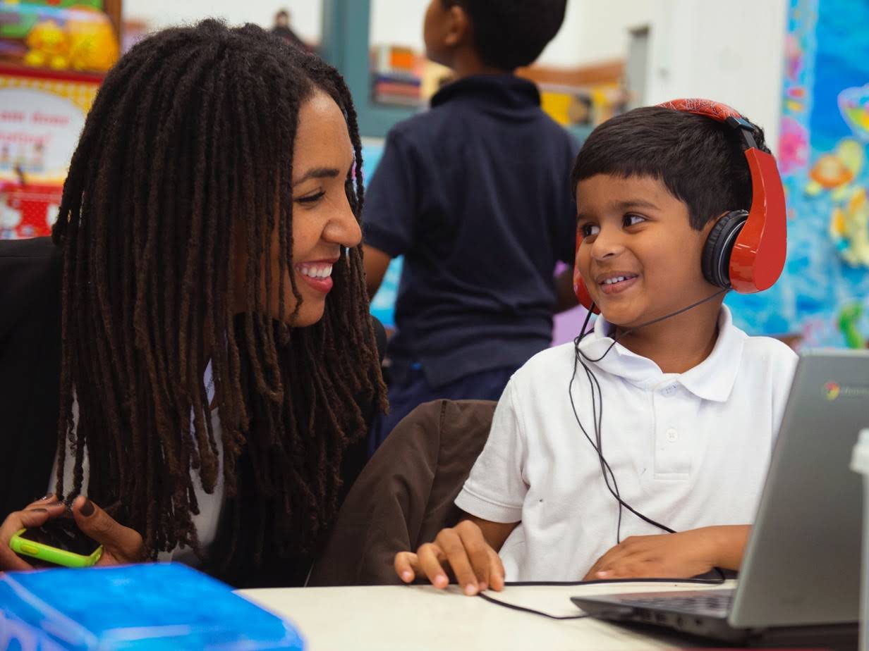 A woman leans over and smiles at a student listening on headphones with a Chromebook in use.