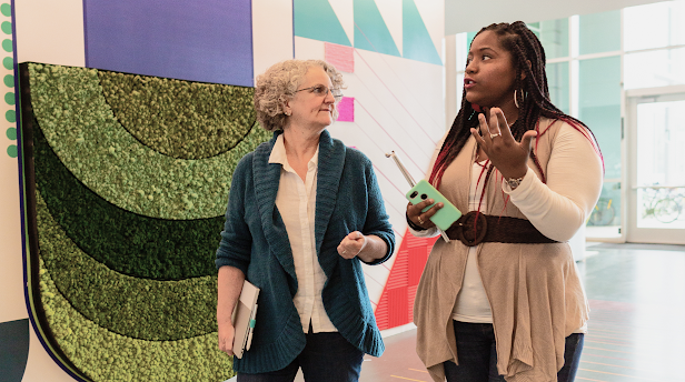 A Black woman with long braids and an older White woman with short gray hair walk engage in conversation as they walk through a hallway with geometric patterns on the wall