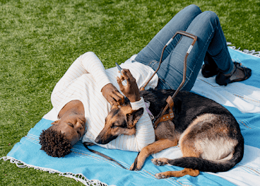 Eine Frau liegt mit ihrem Assistenzhund, einem Schäferhund, auf einer Decke im Gras und hält ein Android-Smartphone in der Hand.