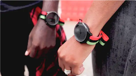 A man and woman wear matching Talley & Twine watches. The watches have black faces and bands striped with red, black, and green.