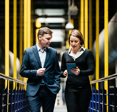 A white man in a blue suit walking next to a white woman in a black sweater reviewing data on a mobile tablet while walking through a data center.
