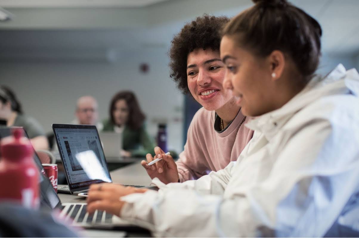 A teenager types on a computer while an instructor engages in providing advice next to her
