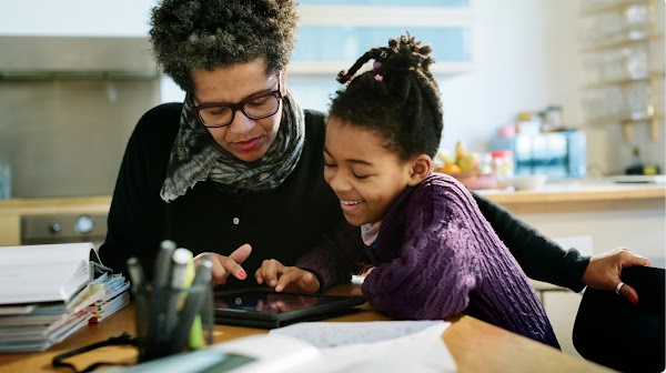 A woman with afro hair and glasses sits and a young girl in braids look at a tablet together.
