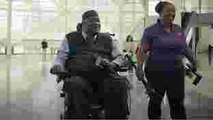Un uomo su una sedia a rotelle che sorride e tiene in mano un microfono e il suo telefono sul ginocchio con una donna in piedi accanto a lui.