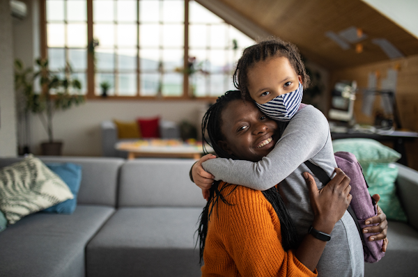 In a living room space, a Black mother warmly smiles and hugs her child