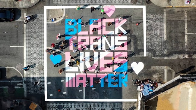 Black Trans Lives Matters mural spray painted in light blue, pink and white in street intersection.