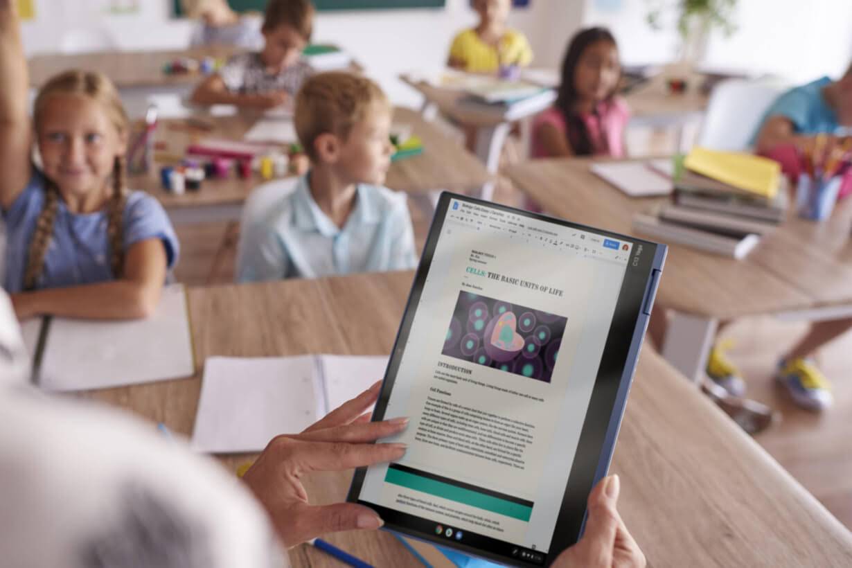 A teacher reads from a Chromebook displaying a document to an attentive class of children.