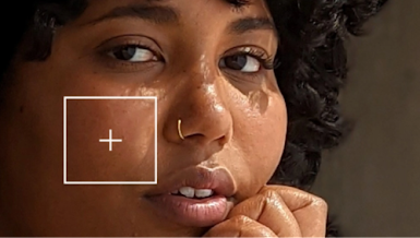 A Black woman with curly hair and freckles stares forward, a framed plus sign is superimposed on her cheek indicating an AI tool analyzing her skin tone