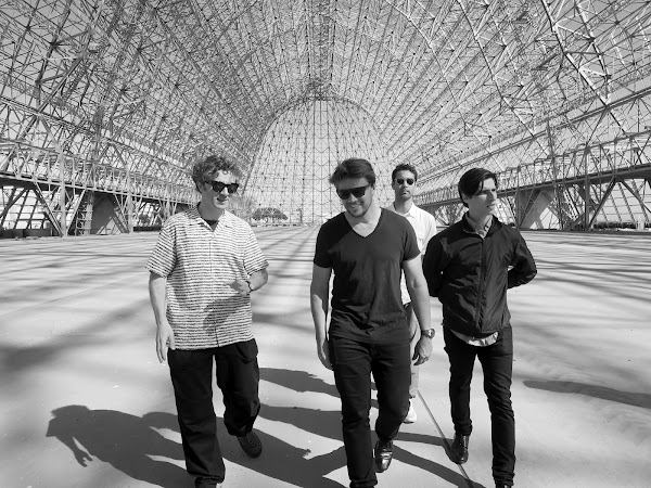 Four men walking together in one direction inside of a very large metal structure