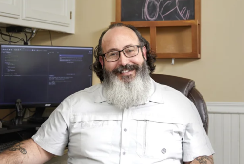Michael Pollack, a bearded man in a white button up shirt and Veteran graduate of Google Career Certificates, sits proudly smiling at his desk.