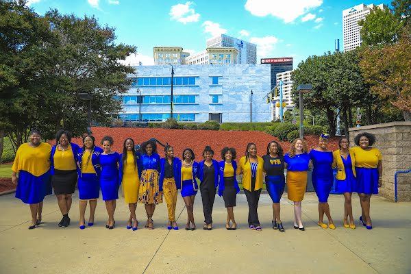 A group of sixteen young Black women standing arm in arm in different blue and yellow professional outfits on a university campus.