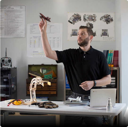 A male teacher stands behind his classroom desk, holding a model airplane in the air with one hand.