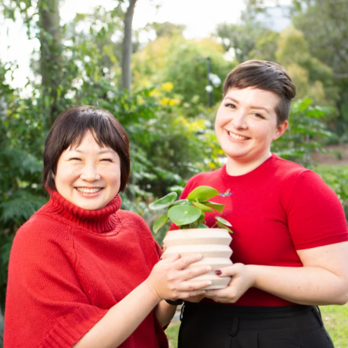 Two women in red tops smile beamingly, holding up a small potted plant together
