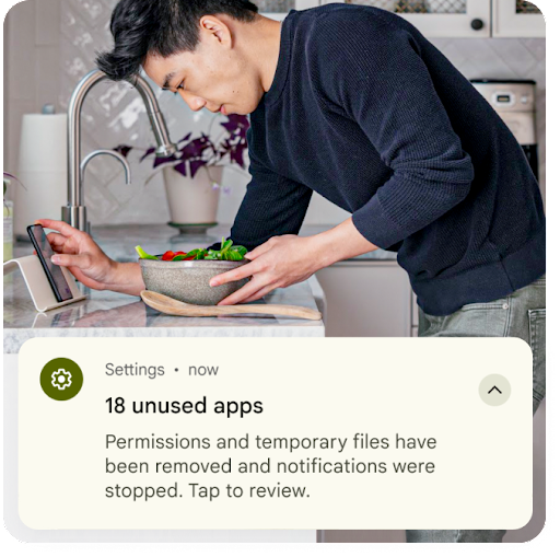 一个人在厨房水槽旁准备食物，同时看着 Android 手机。图片上方的叠加图像显示的是设置通知，指出未使用的应用中的临时文件已被移除，并且已重置权限。