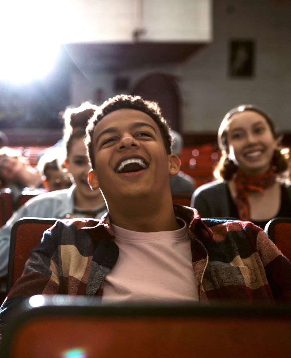 A teenage boy laughs among friends while sitting in a movie theater.