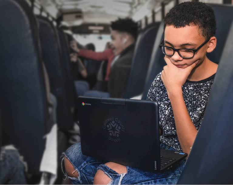A student wearing glasses, seated, focused on a chromebook device in a bus during commute