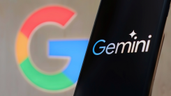 A photograph of a phone displaying the Gemini logo, with the Google logo out of focus against a wall behind the phone